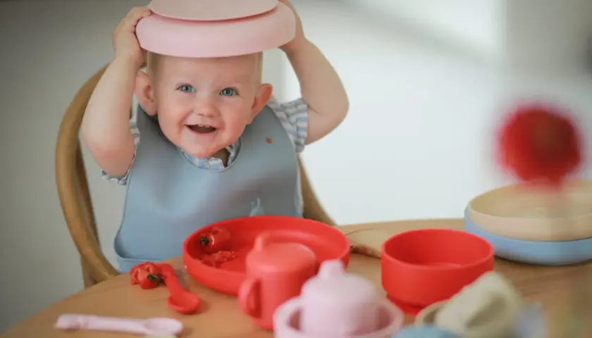 A smiling baby with a pink plate on its head sits at a table with baby feeding dishes.