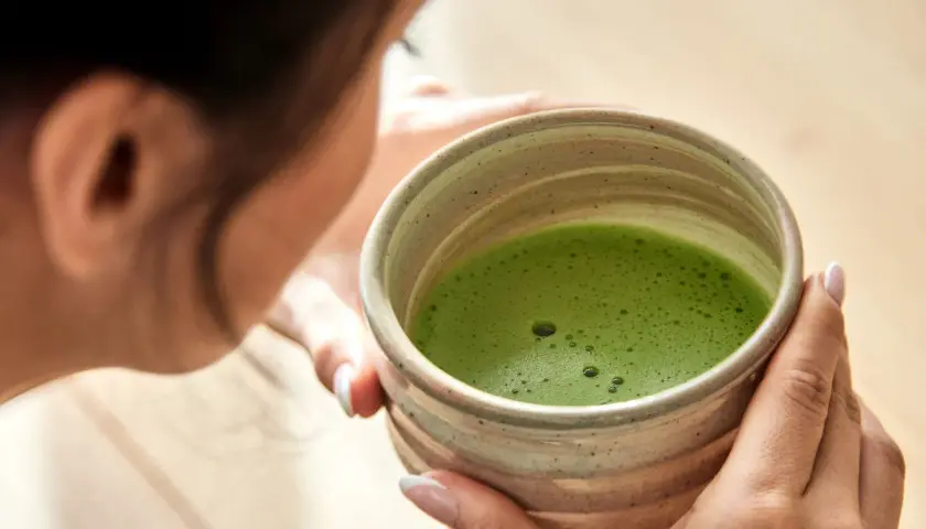 Frau genießt die Matcha Zubereitung in einer traditionellen Schale. Das leuchtend grüne Getränk schäumt leicht.