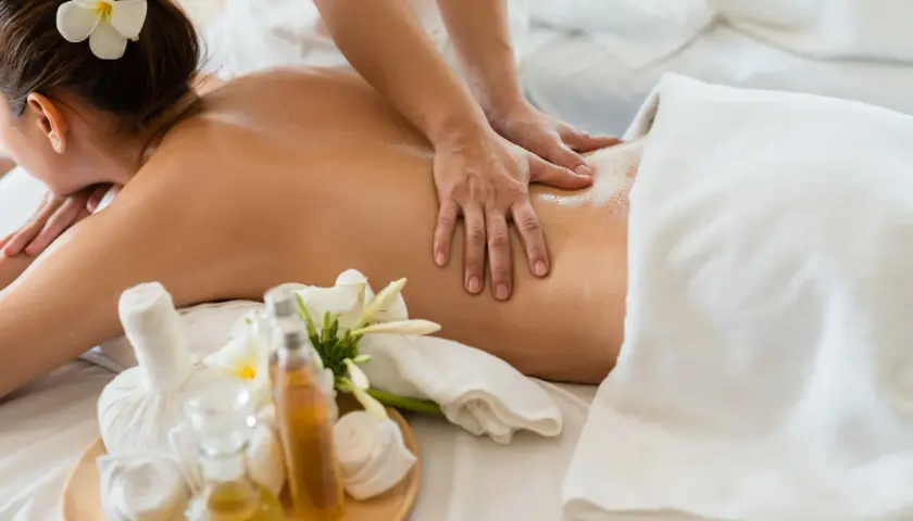 Mains expertes prodiguant un massage américain relaxant sur le dos huilé d'une femme, avec des fleurs de frangipanier et des huiles de massage à proximité.