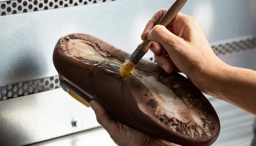 Un cordonnier applique de la colle sur la semelle d'une chaussure marron. Oui, on peut ressemeler des baskets, c'est un art !