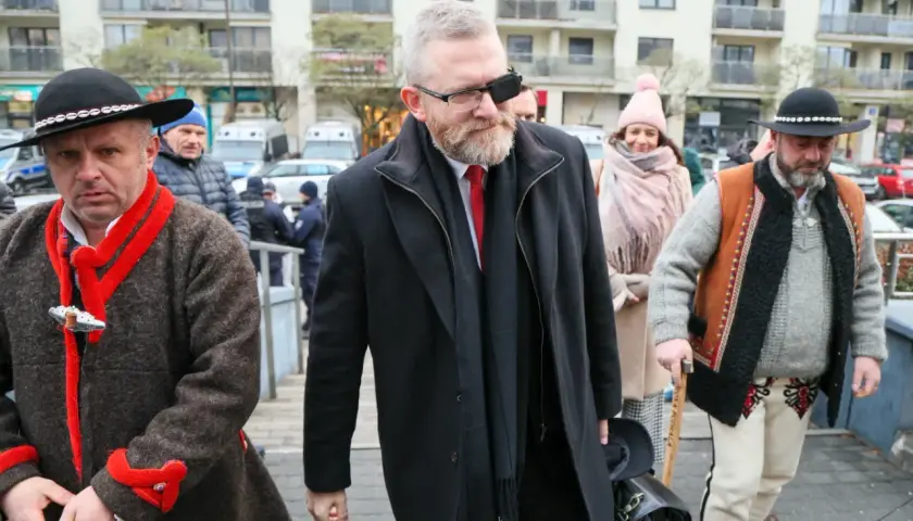 Grzegorz Braun w czarnym płaszczu i z opaską na oku, w otoczeniu mężczyzn w tradycyjnych strojach.