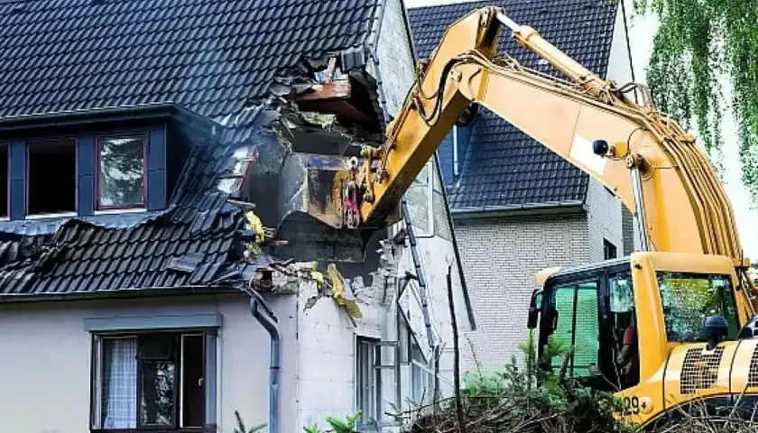 Żółta koparka prowadzi rozbiórkę dachu domu, niszcząc jego konstrukcję.
