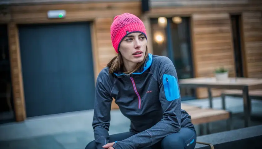 Kobieta w sportowym stroju, z jaskrawym, różowym czapka beanie, siedzi na ławce przed nowoczesnym budynkiem.