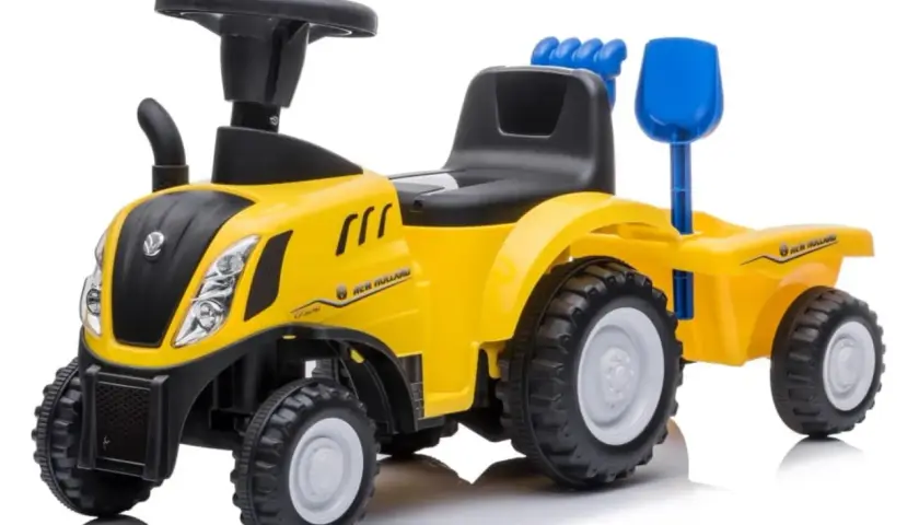 Żółty traktor New Holland z przyczepą, zabawka dla małego rolnika. Ma czarne siedzenie, kierownicę i niebieską łopatkę.