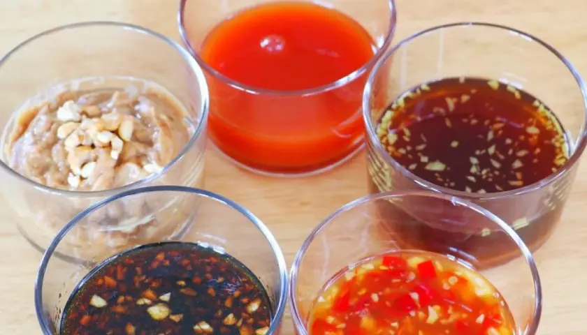 Five small glasses hold various Japanese dipping sauces, including a creamy peanut sauce, a bright red sauce, and a dark soy-based sauce with garlic.