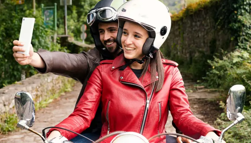 Pareja en moto, él con casco y gafas, ella con casco blanco y chaqueta roja, toman una selfie con el móvil. ¡Manos libres, casco moto legal y a disfrutar del viaje!