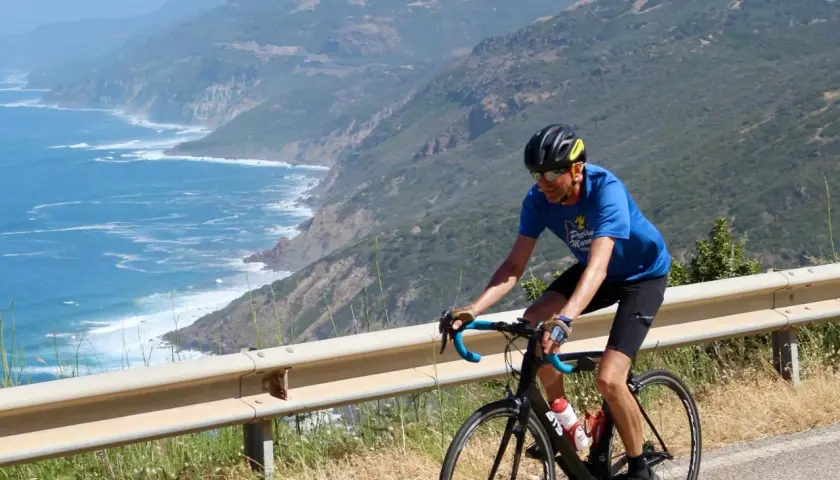 Man on a bike tour in Sardinia, cycling along a scenic coastal road with the blue sea and green hills.