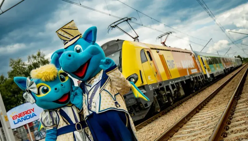 Dwa niebieskie smoki w strojach pozują przy żółtym pociągu. To pociąg do Energylandii, gotowy na podróż pełną wrażeń!