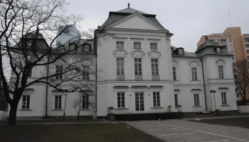 Pałac Przebendowskich, biała, elegancka budowla z zielonym dachem, otoczona drzewami i nowoczesnymi budynkami.
