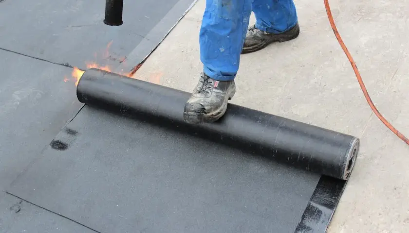 Arbeiter schweißt Bitumenbahn auf Garage Betondecke. Flammen erhitzen die Rolle für die Abdichtung.