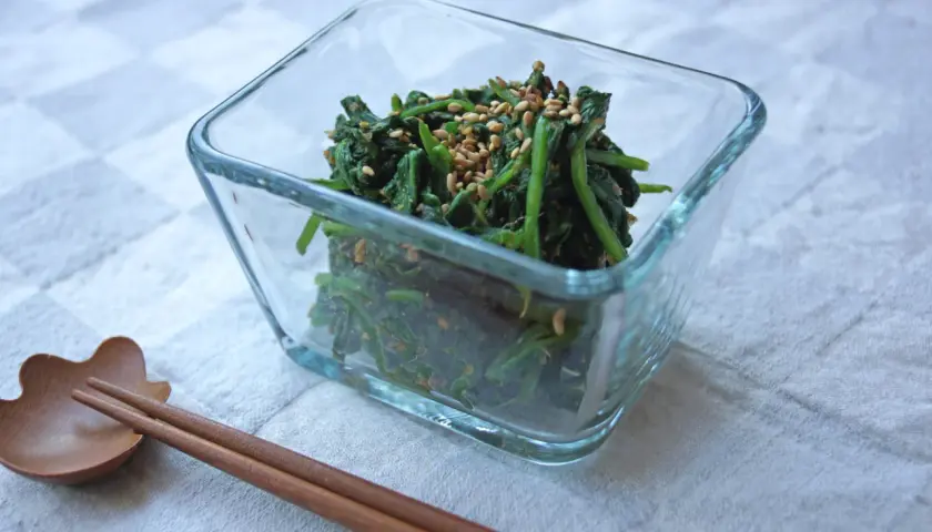 A glass bowl filled with spinach gomae, a savory sesame-dressed spinach dish, is ready to be enjoyed with chopsticks.