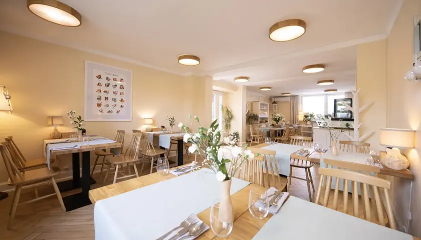Przytulna restauracja z drewnianymi stołami i krzesłami, idealna na urodziny w łodzi dla dorosłych. Jasne ściany, subtelne oświetlenie i świeże kwiaty tworzą miłą atmosferę.