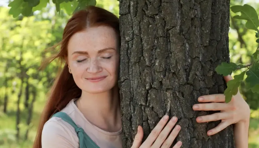 Kobieta z rudymi włosami przytula drzewo. Jej skóra jest pełna piegów, a oczy zamknięte w błogim uśmiechu. To jak naturalne prebiotyki w kosmetykach, które odżywiają i pielęgnują.