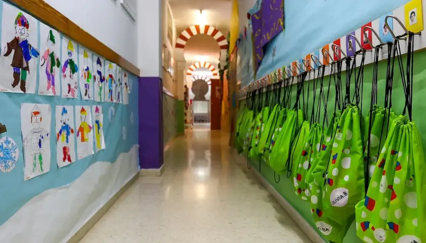 Pasillo de guardería en Comunidad Valenciana. Dibujos infantiles decoran la pared, y mochilas verdes cuelgan listas para la ayuda.