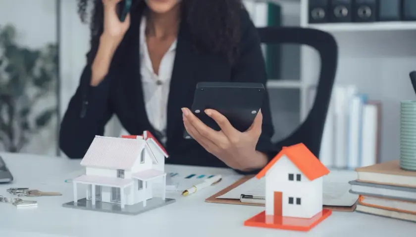Agent immobilier au téléphone, calculatrice en main, devant des maquettes de maisons. Elle prépare une vente à terme.