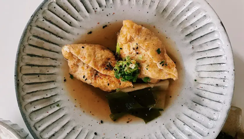 Two pieces of golden aburaage tofu simmer in a savory broth with leeks and seaweed, garnished with green onions and sesame seeds.
