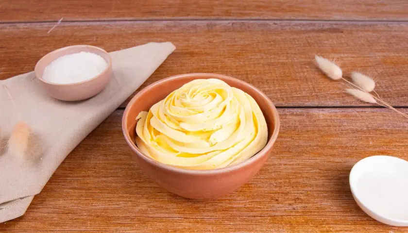 Una ciotola piena di soffice crema al burro, pronta per decorare torte. Accanto, ciotoline con zucchero e decorazioni naturali.