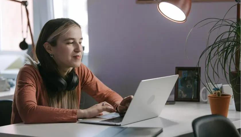 Młoda kobieta z słuchawkami na szyi pracuje na laptopie, być może instaluje aplikację z nieznanych źródeł, ryzykując bezpieczeństwo danych.
