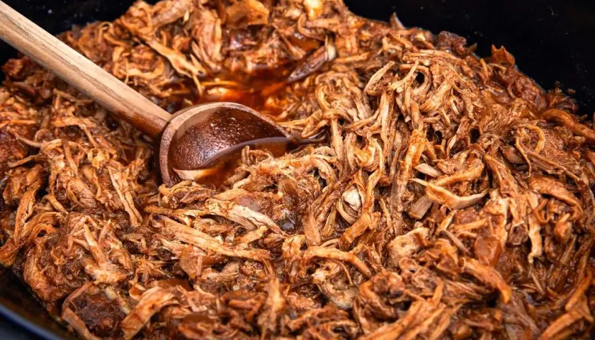 En stor gryta med saftig, långkokt pulled pork i en mörk sås. En träsked vilar i den läckra pulled pork grytan.