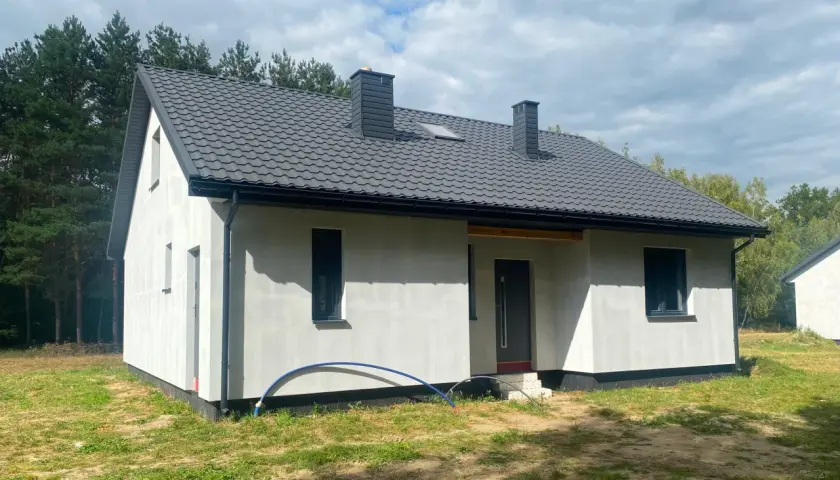 Nowy dom szkieletowy na lata, z szarym dachem i białymi ścianami, otoczony zielenią i lasem.