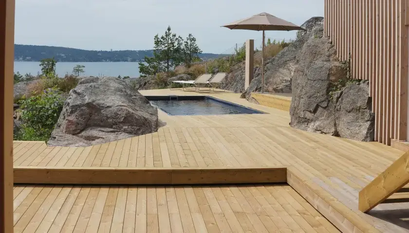 En stor altan i trä med pool och solstolar, omgiven av klippor och med utsikt över vattnet. Trall mått är perfekt för detta sommarparadis.