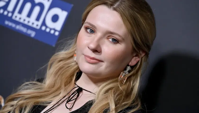 Abigail Breslin, znana z wielu filmów, z niebieskimi oczami i blond włosami, pozuje z uśmiechem.