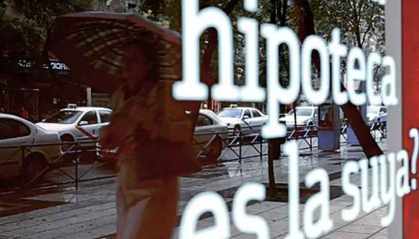 Reflejo en un escaparate: "Hipoteca, ¿es la fein suya?". Una persona con paraguas pasa por la calle lluviosa con taxis.