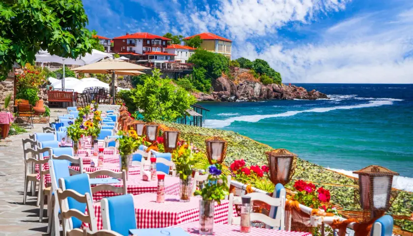 Urokliwa restauracja nad morzem w Bułgarii. Stoliki z czerwono-białymi obrusami i niebieskimi krzesłami czekają na gości, a w tle widać turkusowe morze i malownicze klify.
