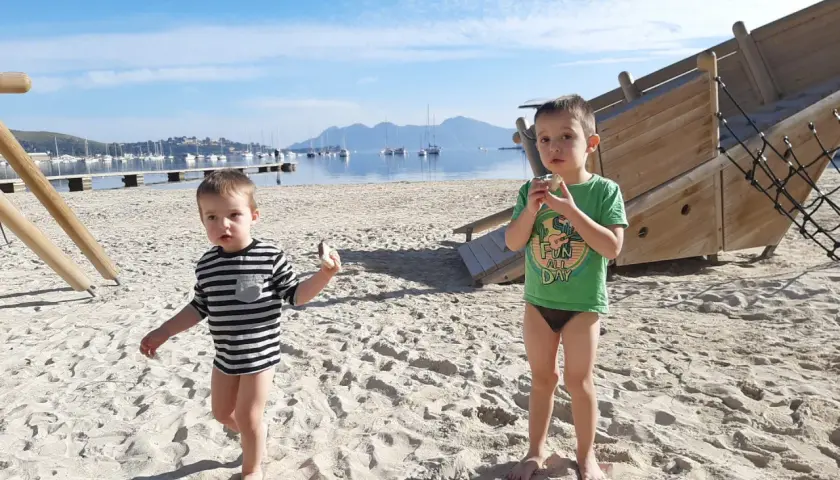 Dwóch chłopców na plaży, cieszących się słoneczną, majową pogodą w lutym. W tle widać morze z łodziami i góry.