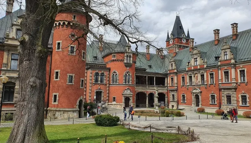 Pławniowice atrakcje: ceglany zamek z wieżą i zegarem, dziedziniec z drzewem i ludźmi spacerującymi po okolicy.