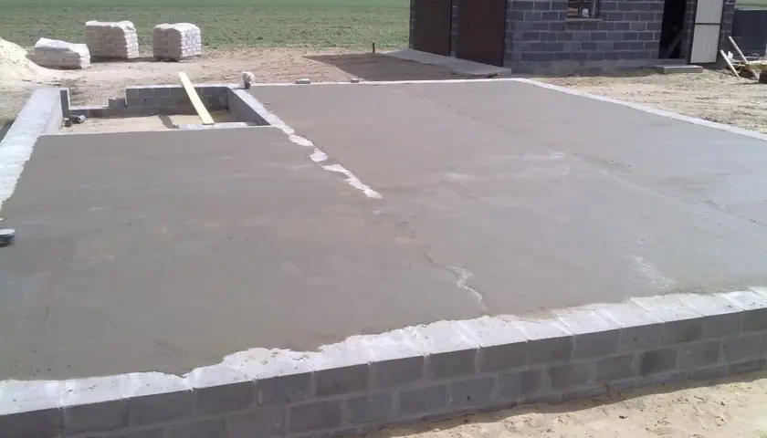 Świeżo wylana płyta betonowa, z widoczną minimalną grubością chudziaka, na fundamencie z bloczków. W tle budynek gospodarczy.