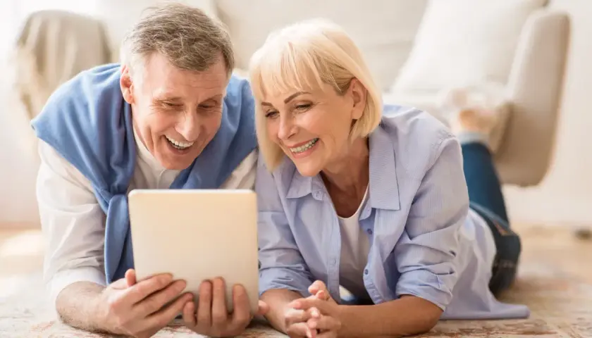 Ein lachendes älteres Paar liegt auf dem Boden und schaut auf ein Tablet. Ihre positiven senioren dates erfahrungen werden durch ihr Lächeln deutlich.