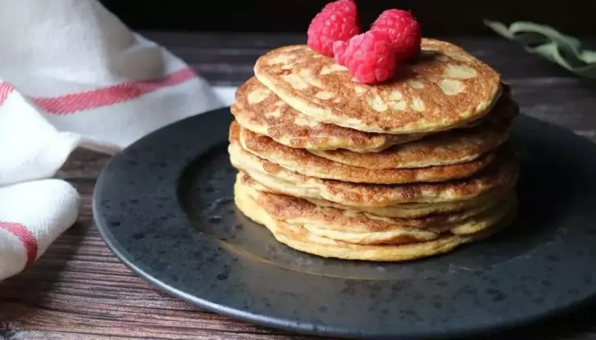 En hög med nyttiga pannkakor toppas med hallon, serverade på en mörk tallrik med en vit handduk med röda ränder i bakgrunden.