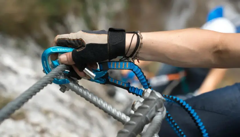 Ręka w rękawiczce z karabinkiem i lonżą, kluczowy element via ferrata sprzętu, zabezpiecza wspinaczkę.