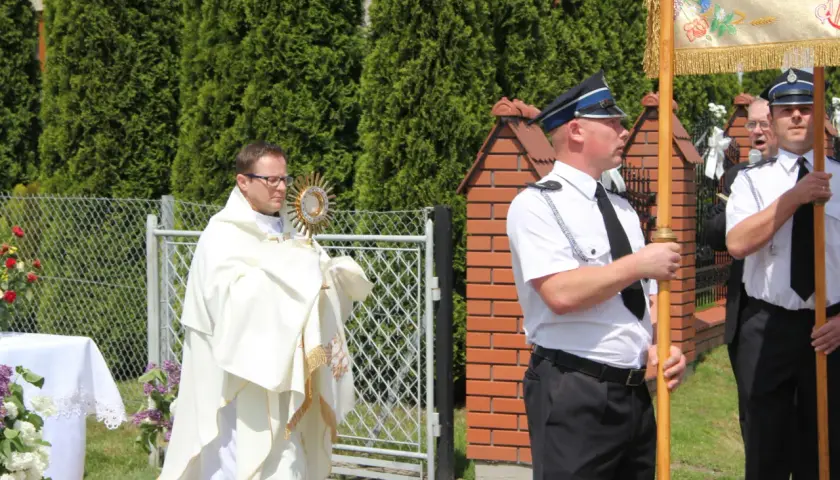 Ksiądz w białej szacie niesie monstrancję, obok stoją mężczyźni w mundurach.