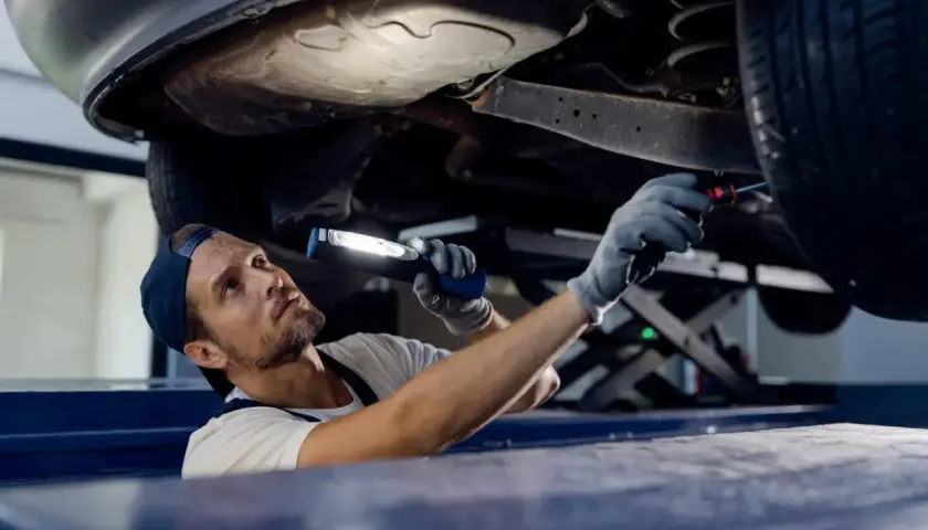 Mechanik z latarką bada podwozie auta. Sprawdza, czy potrzebna jest wymiana wahacza, cena usługi zależy od modelu.