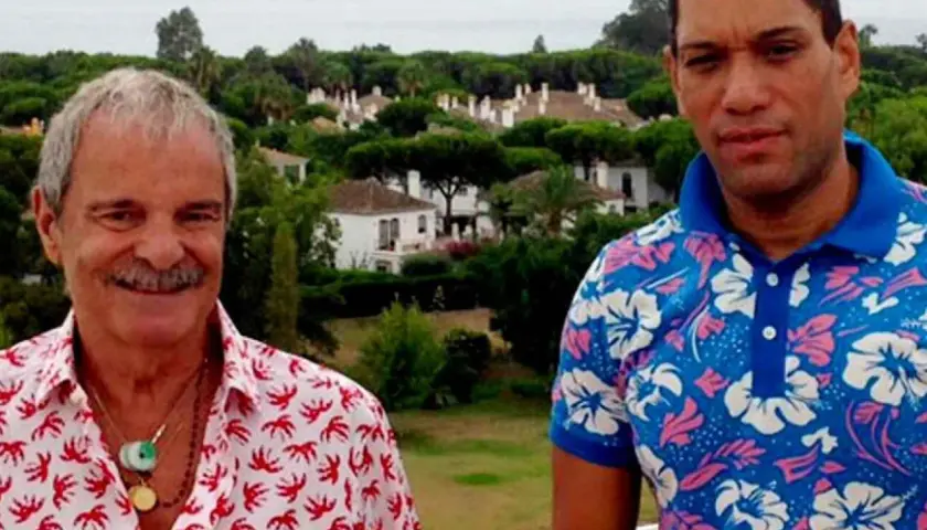 Elio Valderrama Prescott y un amigo posan con camisas coloridas frente a un paisaje de casas y árboles.