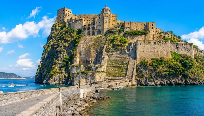The Aragonese Castle on a rocky island, connected by a stone bridge, with boats in the turquoise sea.