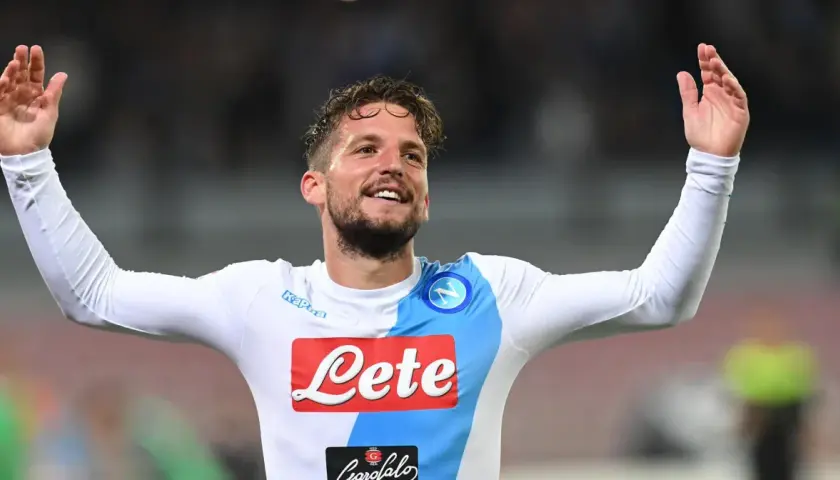 Uśmiechnięty Dries Mertens w białej koszulce Napoli z rękami uniesionymi w geście radości.