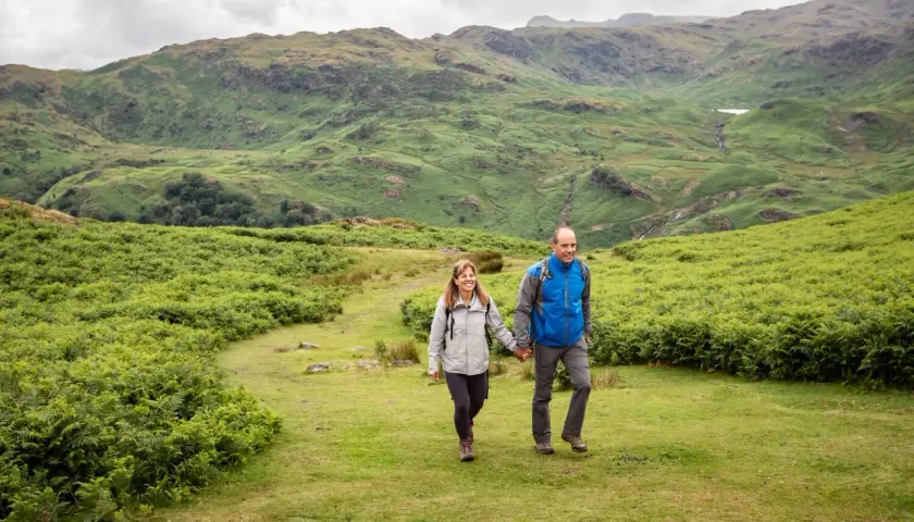 A couple enjoys hiking in England, hand-in-hand, amidst lush green hills and ferns under a cloudy sky.