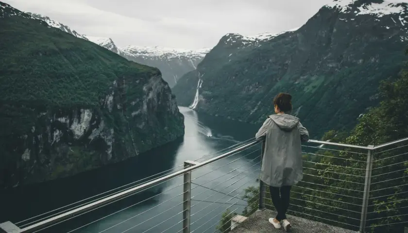 Kobieta podziwia majestatyczne fiordy norweskie, z wodospadem w oddali i ośnieżonymi szczytami gór.