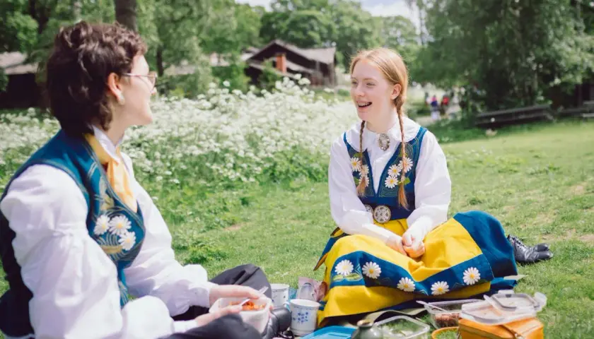 Två personer i folkdräkter njuter av en picknick under midsommar på Skansen.