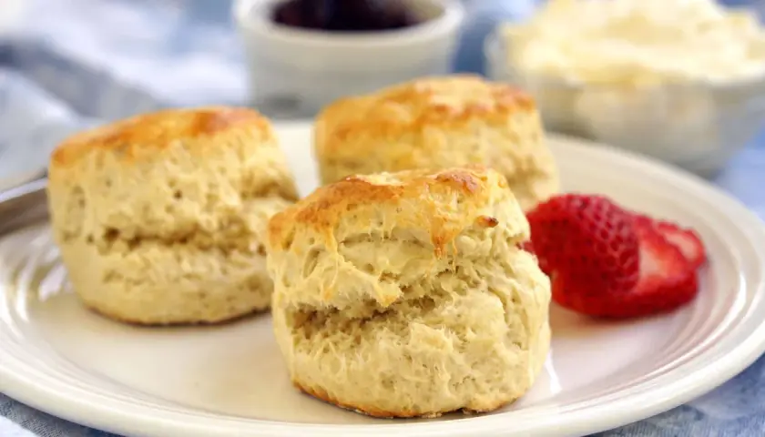 Tre gyllene engelska scones ligger på en vit tallrik med en jordgubbe. I bakgrunden syns sylt och grädde.