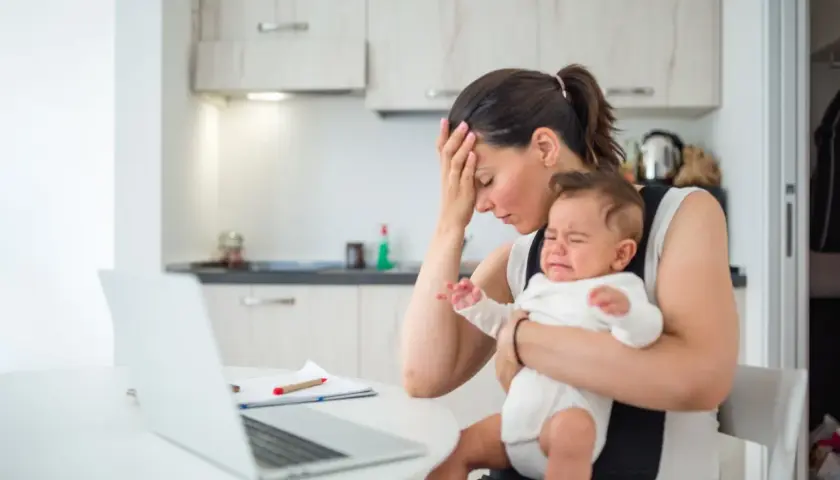 Zmęczona mama z płaczącym, **high need baby** na rękach, próbuje pracować przy laptopie.