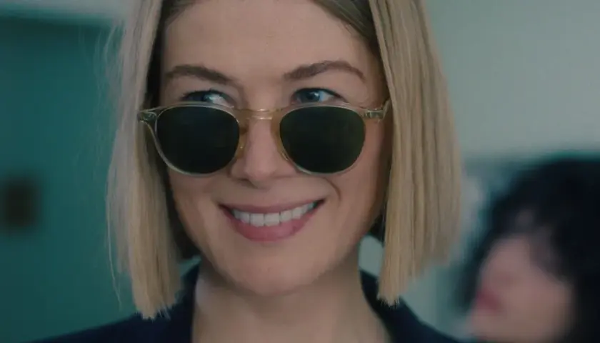 Uśmiechnięta Rosamund Pike w okularach przeciwsłonecznych, przypominająca scenę z jej filmów i seriali.