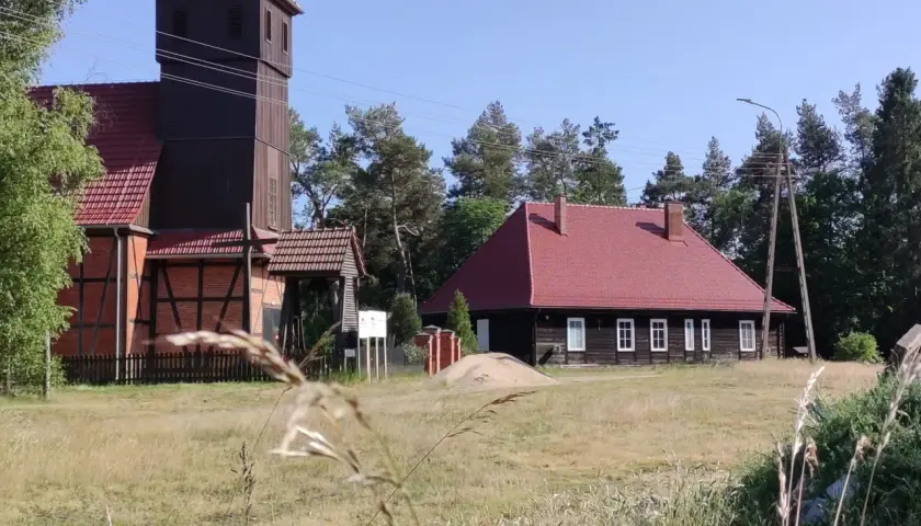 Drewniana wieża kościelna i dom z czerwonym dachem w Starowej Górze, otoczone polami i drzewami.