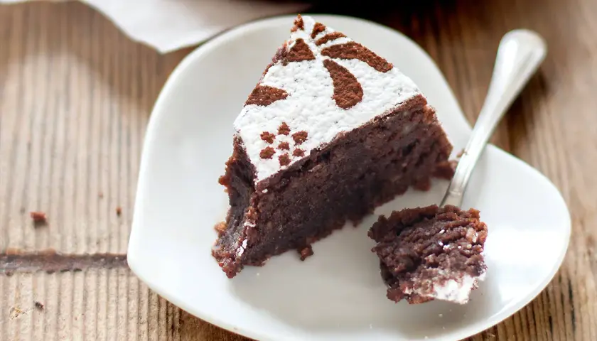 Una fetta di torta caprese ricetta originale napoletana, cosparsa di zucchero a velo e cacao, su un piatto bianco a forma di cuore.