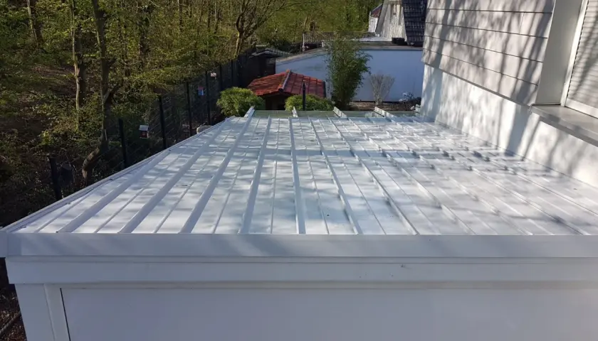 Neues, weißes Metalldach mit sichtbarem Ortgang. Im Hintergrund ein kleiner Garten mit Bäumen und einem roten Gartenhaus.