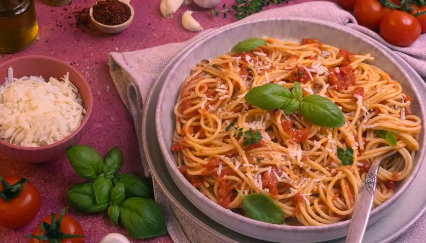 Ein Teller Spaghetti Napoli mit Tomatensauce, Basilikum und Parmesan. Daneben frische Tomaten, Knoblauch und Kräuter.