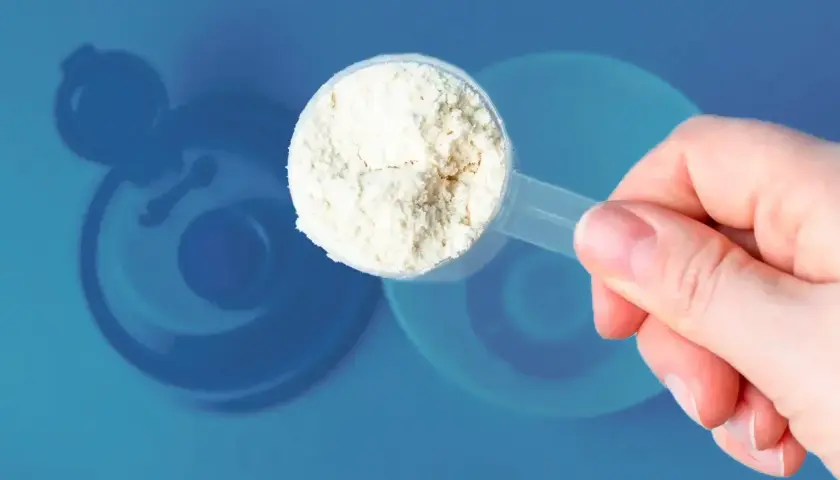 Una mano sostiene una cuchara con polvo blanco, ¿cuanta creatina tomar? Fondo azul con silueta de batidora.